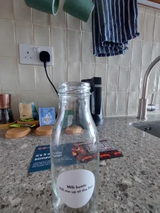 room 2 Chiswick glass milk bottle on a kitchenette counter, labelled “Milk bottle Fill me up at the bar,” with tea and coffee supplies in the background.