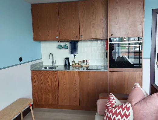 In-room kitchenette at room2 Chiswick hometel in London with wooden cabinets, sink, oven, and compact cooking area beside a sofa for flexible long stays