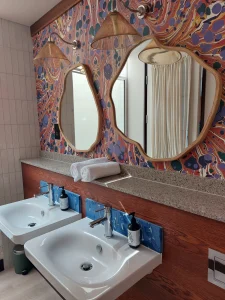 Art-inspired bathroom at room2 Chiswick hometel in London with two sinks, terrazzo countertop, brass wall lights, and colorful marbled wallpaper behind organic-shaped mirrors
