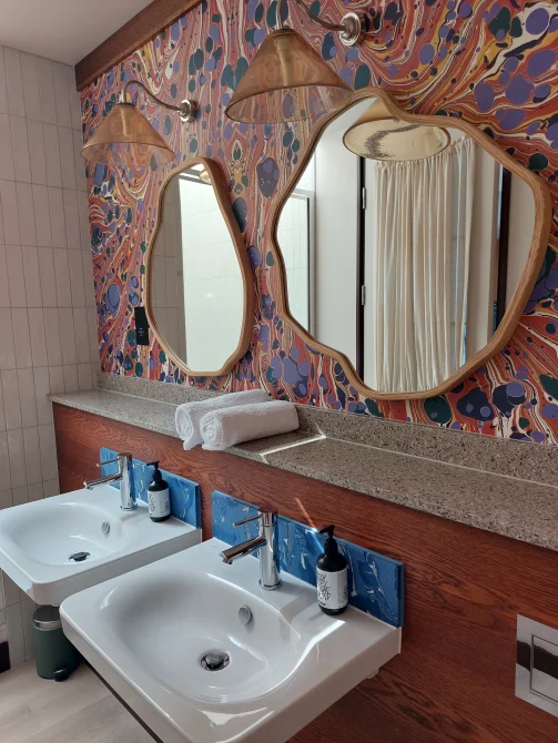 Art-inspired bathroom at room2 Chiswick hometel in London with two sinks, terrazzo countertop, brass wall lights, and colorful marbled wallpaper behind organic-shaped mirrors