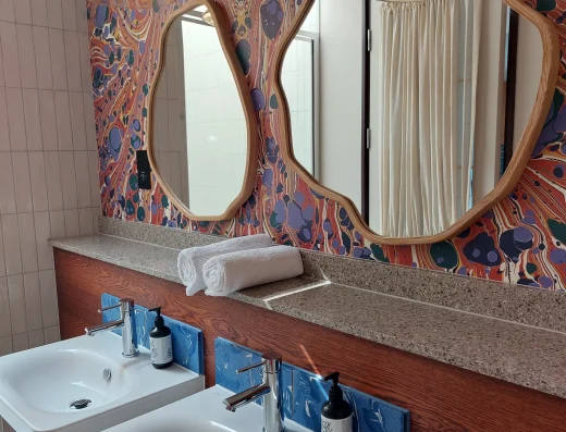 Art-inspired bathroom at room2 Chiswick hometel in London with two sinks, terrazzo countertop, brass wall lights, and colorful marbled wallpaper behind organic-shaped mirrors