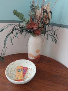 room 2 Chiswick Ceramic vase with dried flowers on a wooden table, with a speckled bowl holding two chocolate bars.