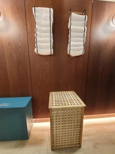 room2 Chiswick fitness room towel station with neatly stacked white towels on wall racks, a wooden laundry basket, and a blue zero-waste bin.