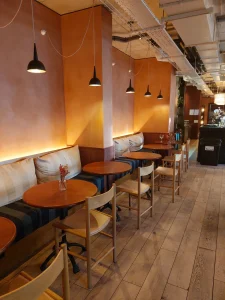 room2 Chiswick café seating with small round tables, wooden chairs, striped banquette seating, and black pendant lights along a warm-toned wall