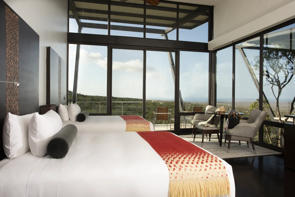 A modern queen bedroom with floor-to-ceiling windows, private balcony, panoramic forest and ocean views, contemporary furnishings, and natural light.