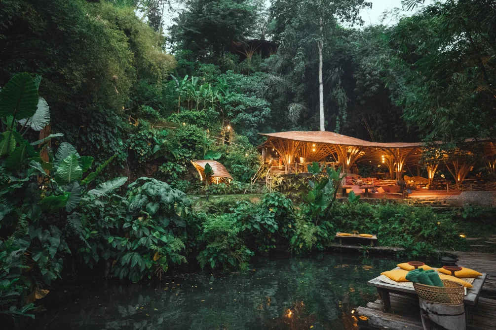 A bamboo pavilion with warm lighting sits beside a dark river pool, surrounded by dense jungle vegetation and tropical plants.