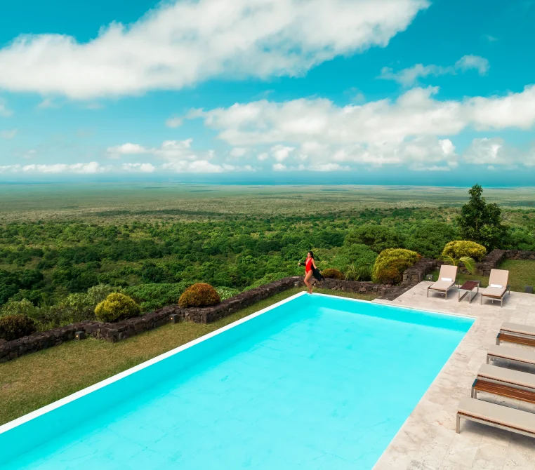 Aerial view of a turquoise pool overlooking vast Galápagos landscape with lush green forest, ocean horizon, and a jogger on the grounds.