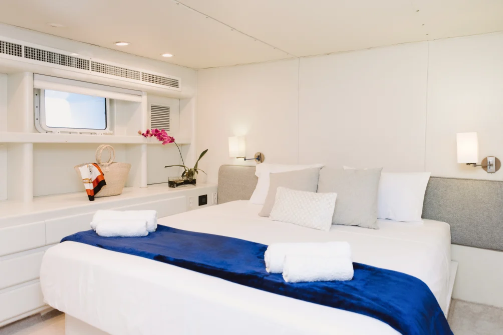 A modern yacht cabin with a queen bed, white linens, blue accent throw, porthole window, built-in desk, and fresh orchid flowers.