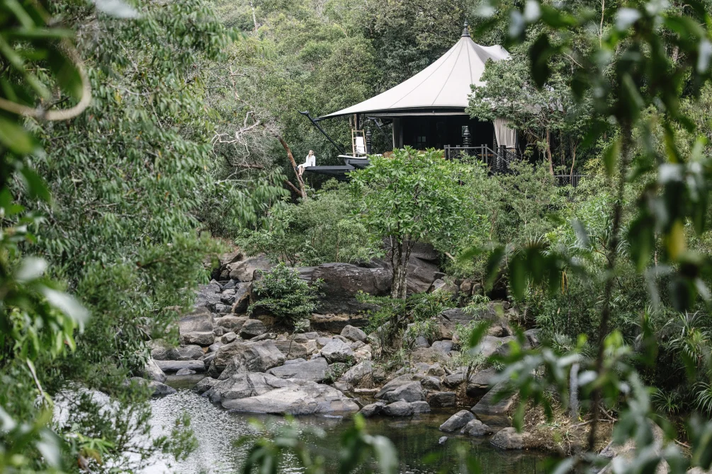 A luxury tented pavilion with cream canvas roof overlooking a rocky stream, framed by lush Cambodian forest vegetation.