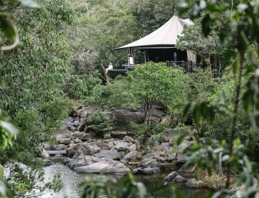 A luxury tented pavilion with cream canvas roof overlooking a rocky stream, framed by lush Cambodian forest vegetation.