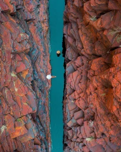 Aerial view of Joffre Gorge at Karijini Eco Retreat, Western Australia, showing turquoise waterhole between red rock cliffs with two swimmers