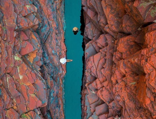 Aerial view of Joffre Gorge at Karijini Eco Retreat, Western Australia, showing turquoise waterhole between red rock cliffs with two swimmers