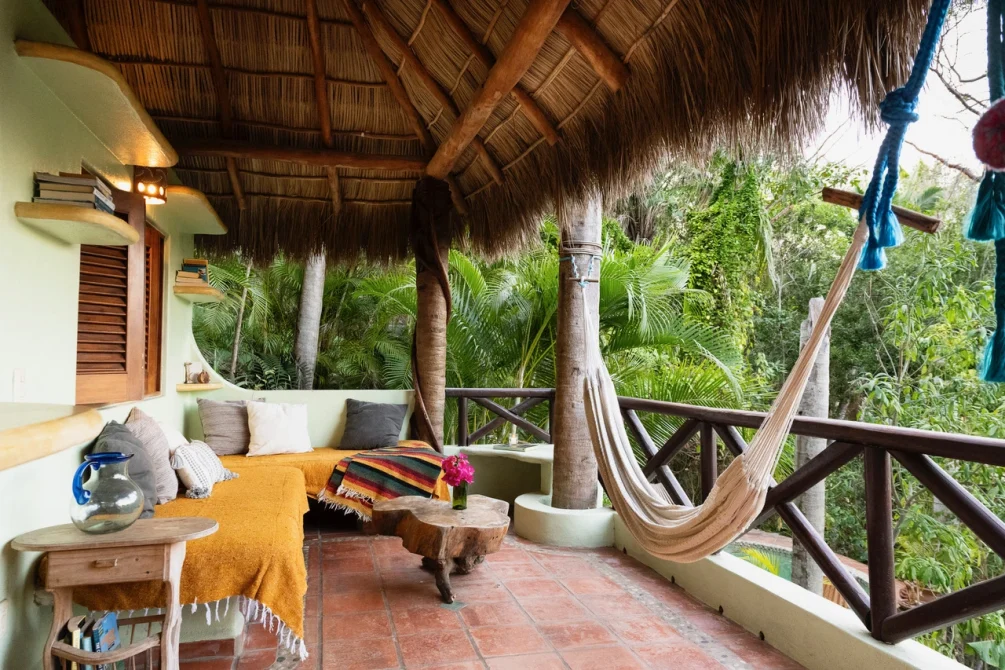 A spacious thatched-roof terrace with a hammock, built-in seating, colorful blankets, and views of dense tropical forest.