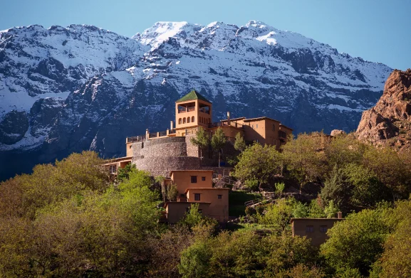 Kasbah du Toubkal perched on hillside with green-roofed tower and snow-capped Atlas Mountains in the background