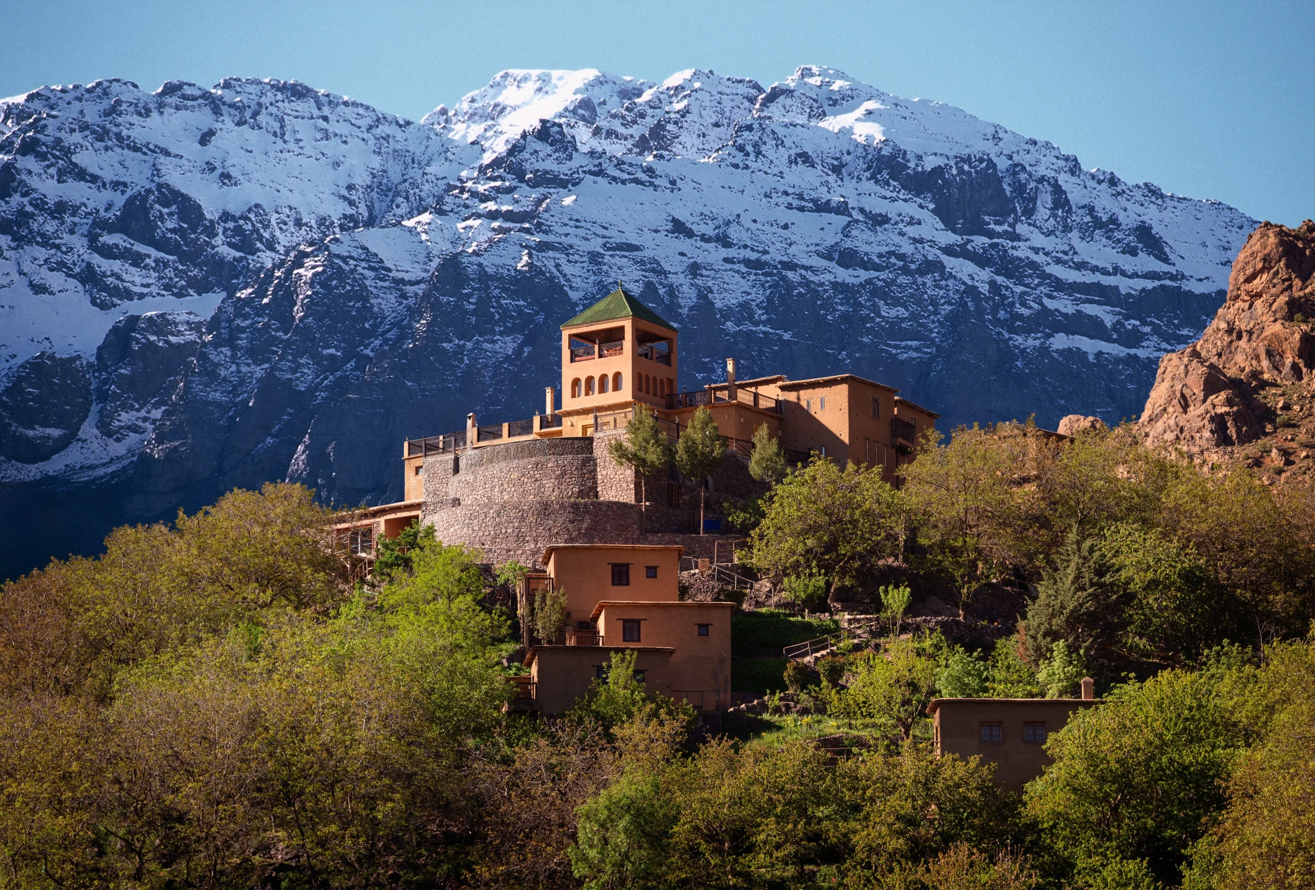 Kasbah du Toubkal perched on hillside with green-roofed tower and snow-capped Atlas Mountains in the background