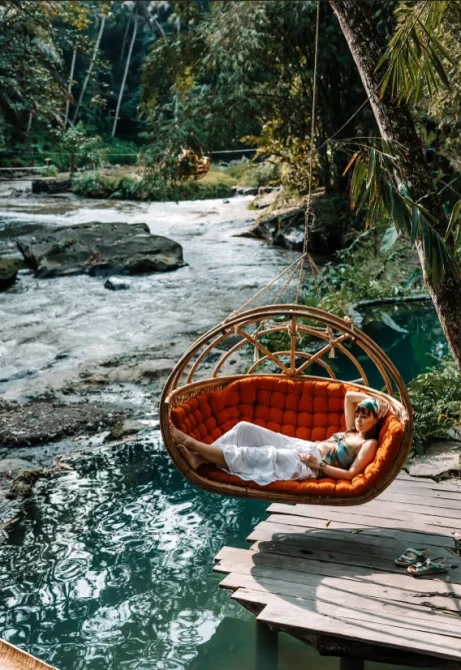 A guest relaxes on a hanging rattan daybed above a clear river pool beside a wooden deck, with lush jungle and flowing water behind.