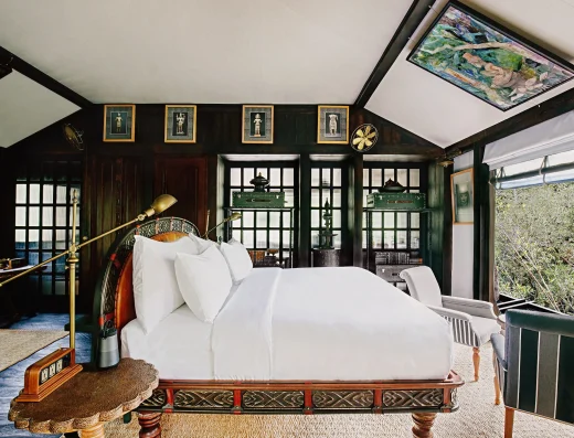 A luxurious bedroom with a carved wooden bed, white linens, brass reading lamp, dark green walls with gold-framed Khmer art, and forest views through glass doors.