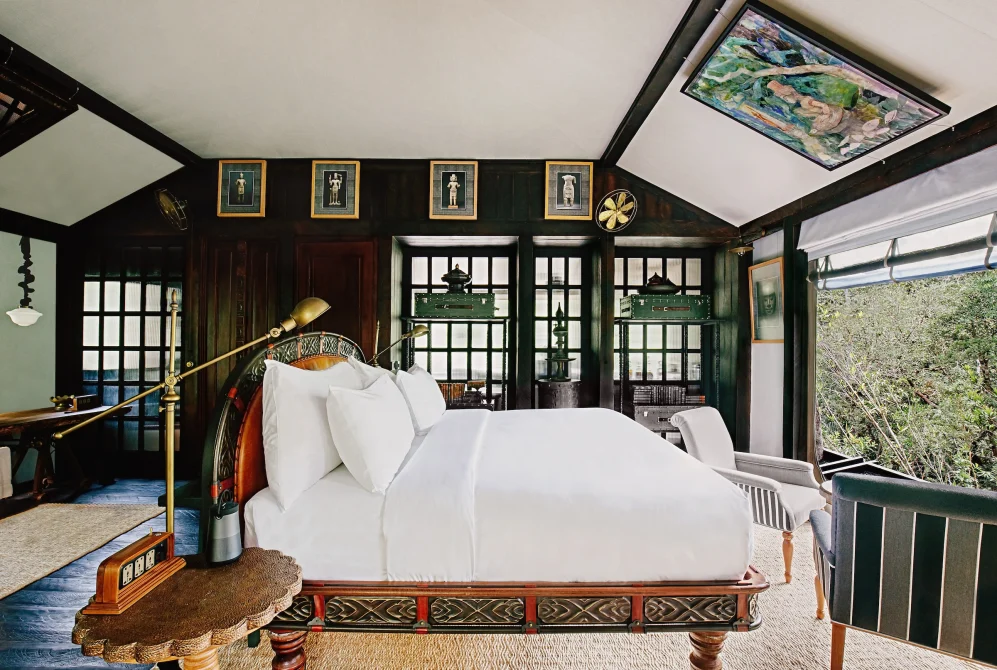 A luxurious bedroom with a carved wooden bed, white linens, brass reading lamp, dark green walls with gold-framed Khmer art, and forest views through glass doors.