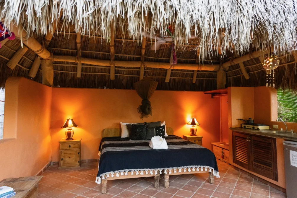 A vibrant orange bedroom with a thatched roof, queen bed with navy bedding, wooden nightstands with warm lighting, and rustic furnishings.