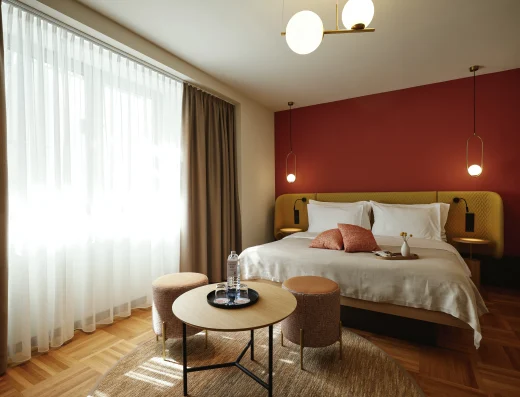 Modern hotel suite at Hotel Henriette Vienna featuring a comfortable bed with mustard headboard, burgundy accent wall, contemporary pendant lighting, and a seating area with round wooden table