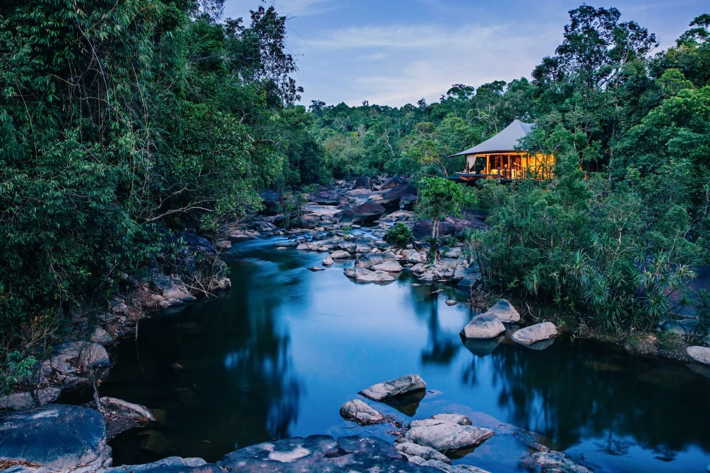 A luxury tented lodge glowing at dusk above a rocky river, surrounded by dense Cambodian rainforest.