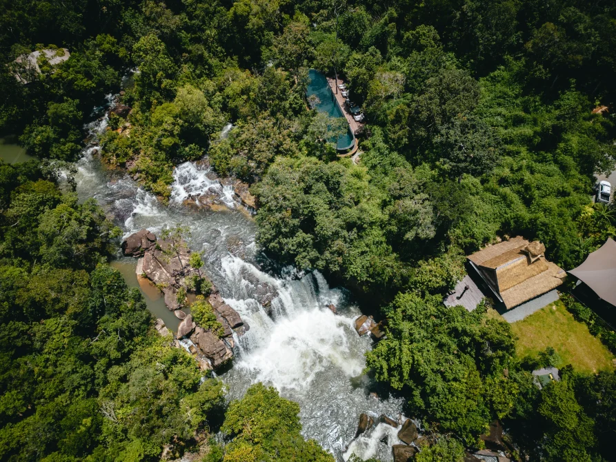 Aerial view of a rainforest river waterfall with a long narrow pool and lodge structures tucked into dense jungle.