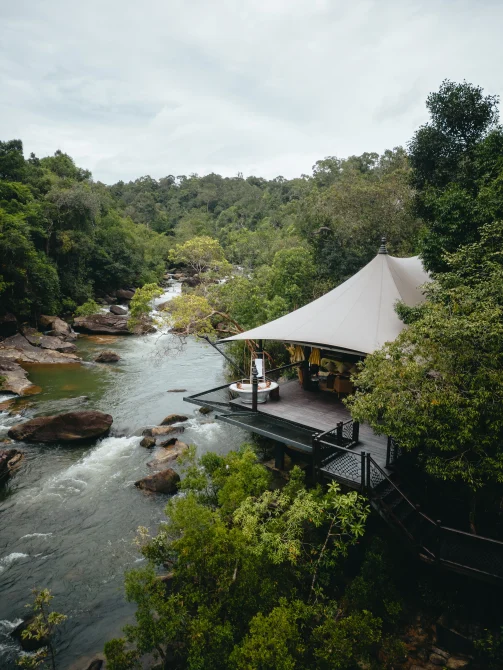 A cream canvas tent pavilion with a dark deck overlooks a rushing river surrounded by lush Cambodian rainforest.