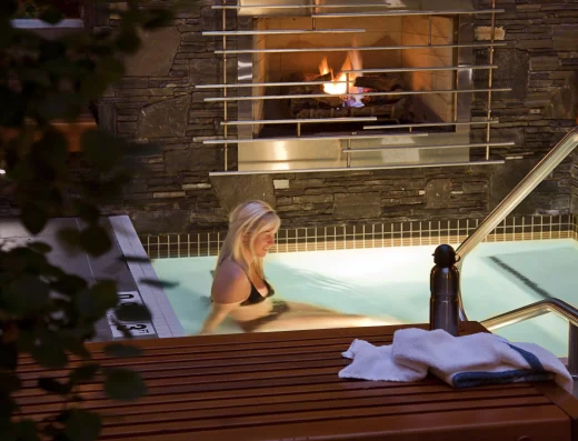 ALT TEXT Outdoor hot tub at Banff Aspen Lodge in Banff, Alberta with a guest soaking in warm water beside a stone fireplace and cozy mountain lodge setting
