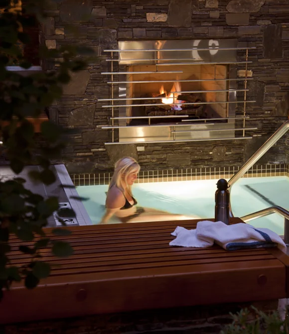 ALT TEXT Outdoor hot tub at Banff Aspen Lodge in Banff, Alberta with a guest soaking in warm water beside a stone fireplace and cozy mountain lodge setting