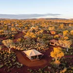 Aerial view of Karijini Eco Retreat with safari tents and cabins scattered across red outback landscape and native bush at sunset