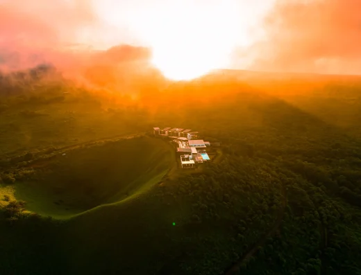 Aerial view of Pikaia Lodge nestled in lush green hills at golden hour sunset, with bungalows, main building, and pool visible among dense vegetation.