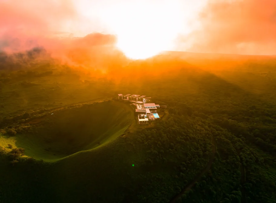 Aerial view of Pikaia Lodge nestled in lush green hills at golden hour sunset, with bungalows, main building, and pool visible among dense vegetation.