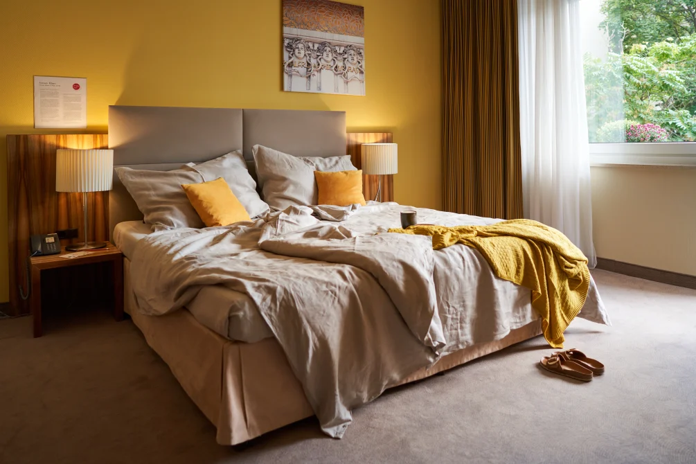 Bright bedroom at Hotel Henriette Vienna with golden yellow walls, comfortable bed with neutral linens and gold accents, wooden nightstands, and large windows overlooking green gardens