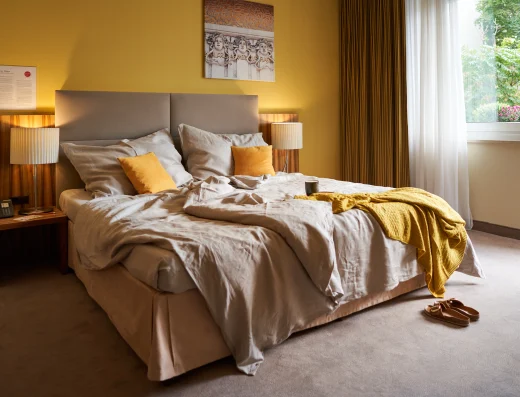 Bright bedroom at Hotel Henriette Vienna with golden yellow walls, comfortable bed with neutral linens and gold accents, wooden nightstands, and large windows overlooking green gardens