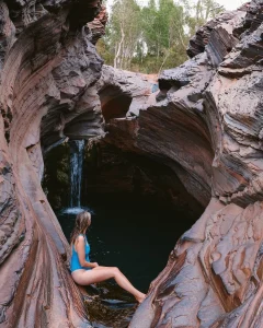 Woman sitting on red rock ledge at a natural waterfall pool inside Karijini gorge, Western Australia, with cascading water and layered rock formations