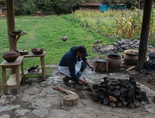 Local guide preparing traditional Andean cooking over an open fire with ceramic pots and farm animals in the background at El Albergue