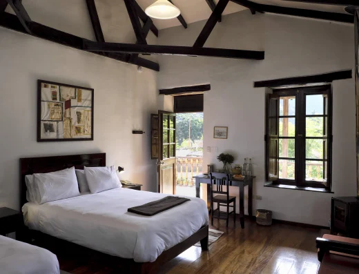 Bright guest bedroom with white linens, dark wooden beams, and large windows overlooking the Sacred Valley at El Albergue