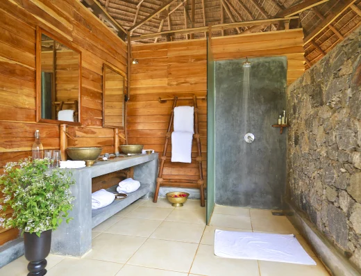 Eco-lodge bathroom with wooden walls, thatched roof, concrete vanity with brass basin, open-air stone shower, potted plant, white towels, and natural materials creating an outdoor bathing sanctuary.