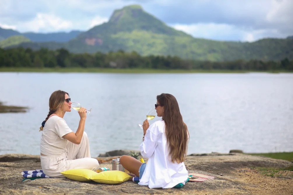Two women sitting on beach with yellow pillow, sipping white wine, overlooking calm lake with forested mountain peak in background.