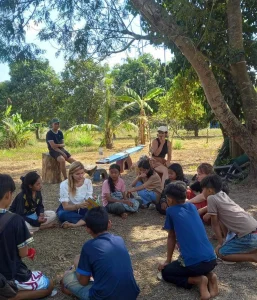 Volunteers and local children sit in a circle under tall trees.