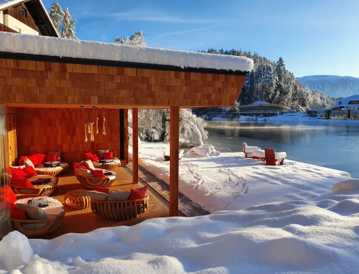A cozy lakeside lounge pavilion with red cushions beside a snowy Alpine lake at Weihrerhof, with winter forest and mountains in the background.