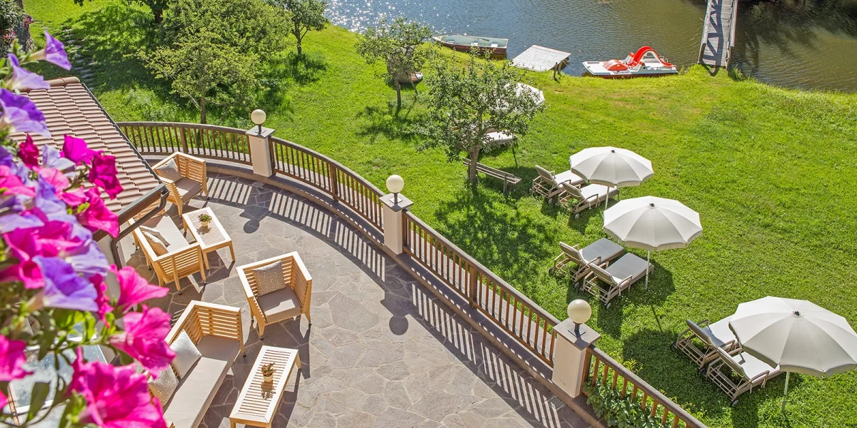 A lakeside terrace at Weihrerhof with wooden loungers, white umbrellas, flowering vines, and boats on the water in summer sunlight.