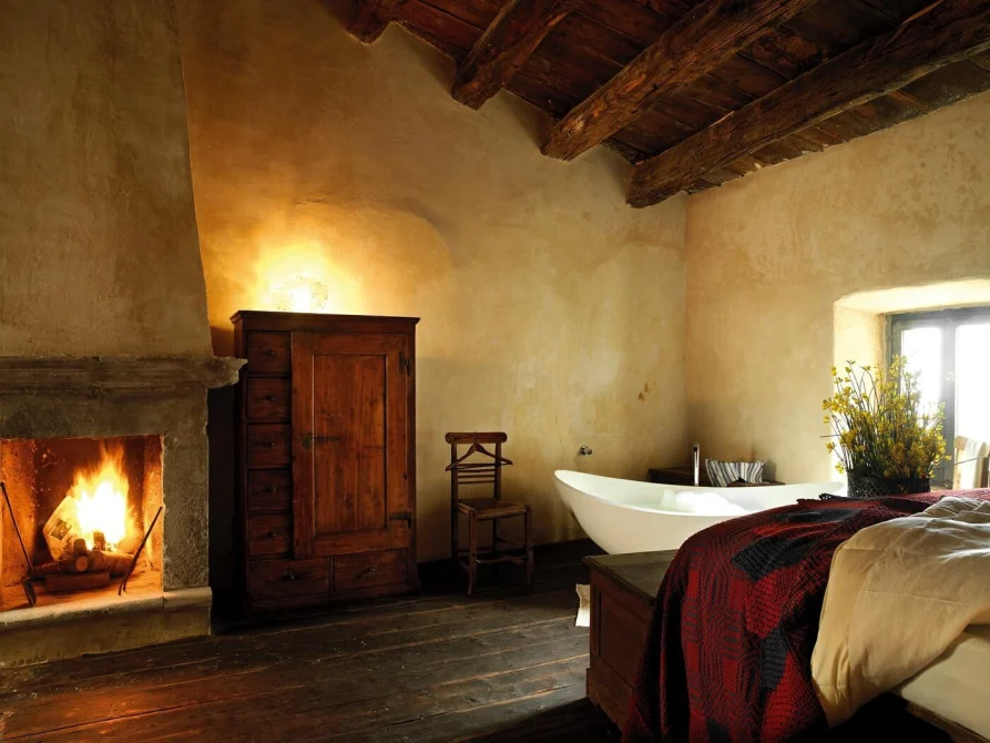 A rustic bedroom with a fireplace, wooden beams, a freestanding soaking tub, antique furniture, and warm candlelit ambiance in a stone village room.