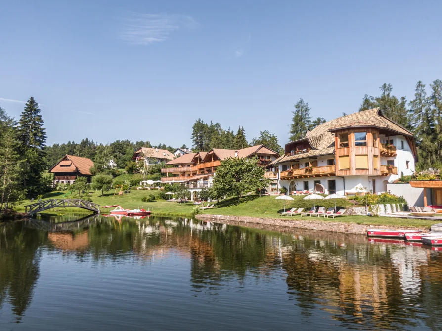 Weihrerhof resort viewed from across the lake, showing traditional Alpine architecture with wooden buildings, manicured lawns, and red boats moored at the shore.