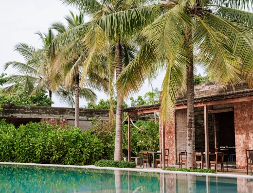 Tropical pool with palm trees and a terracotta stone building featuring an open-air dining area beside the water.