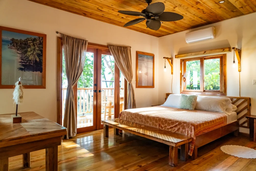 A bright wooden bedroom with a queen bed, ceiling fan, balcony doors, and tropical views at Casa Cayuco in Bocas del Toro, Panama.