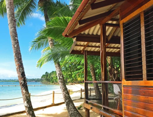 A beachfront bungalow with wooden construction and louvered windows sits between palm trees on white sand, facing turquoise Caribbean waters at Casa Cayuco in Bocas del Toro, Panama.