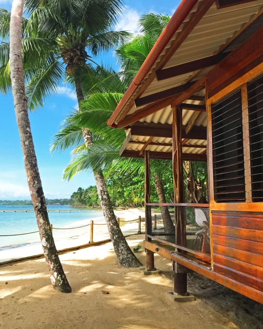 A beachfront bungalow with wooden construction and louvered windows sits between palm trees on white sand, facing turquoise Caribbean waters at Casa Cayuco in Bocas del Toro, Panama.