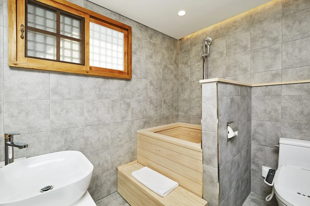 A modern hanok bathroom blending traditional wooden soaking tub with contemporary fixtures, stone tiles, and latticed window.