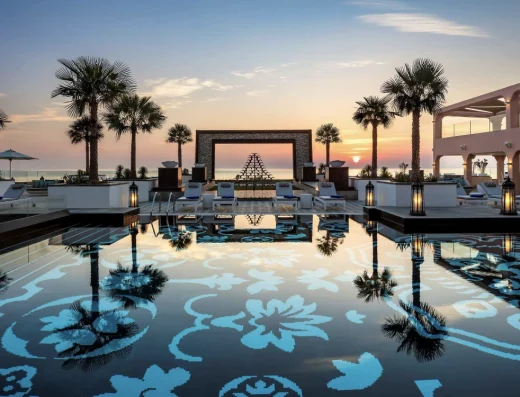 A luxurious resort pool at sunset with ornate blue mosaic patterns, palm trees, architectural frame, and Gulf Sea views glowing in golden light.